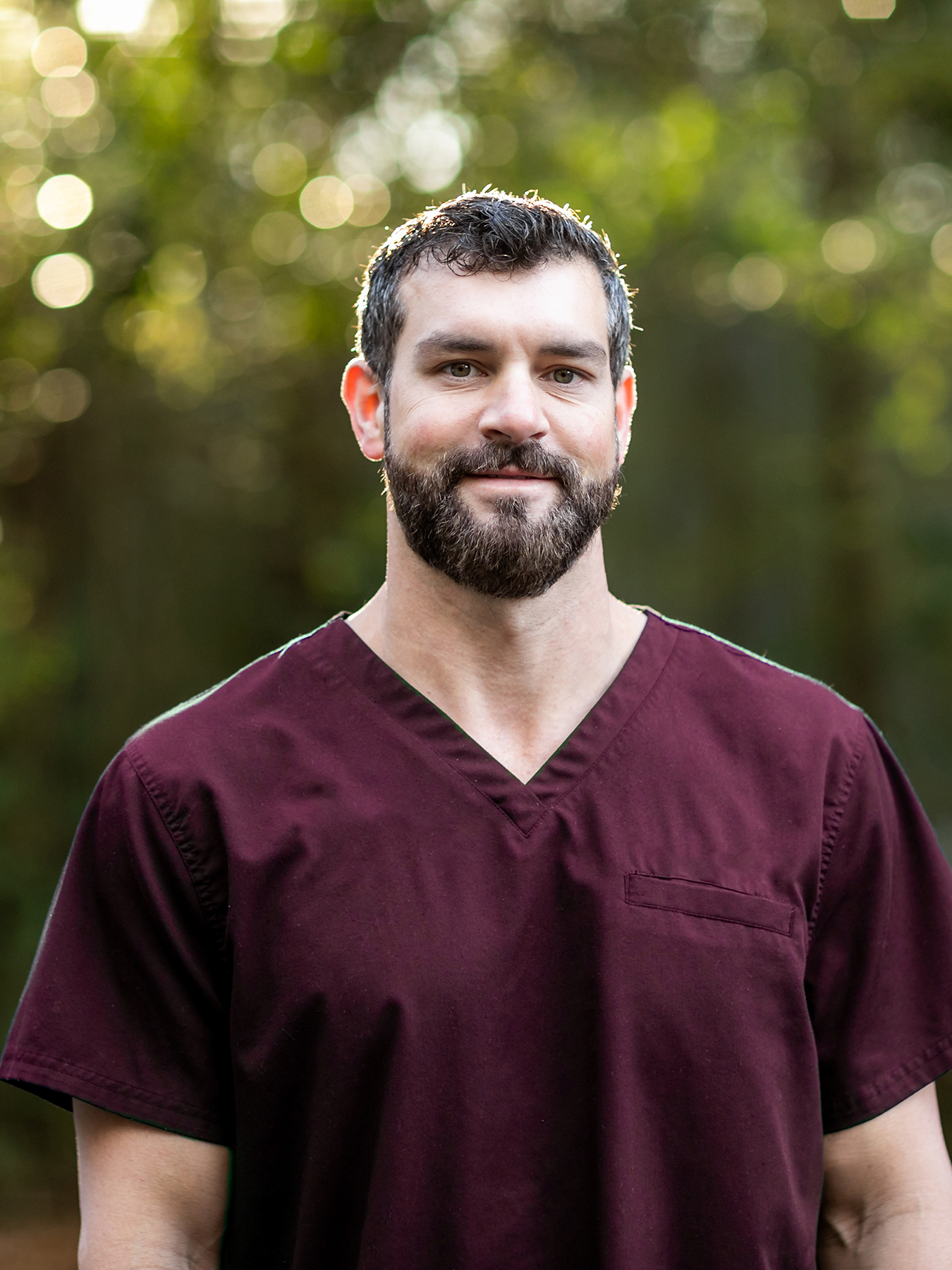 Image of a doctor with dark hair and a beard in maroon scrubs standing outside in a natural green environment.