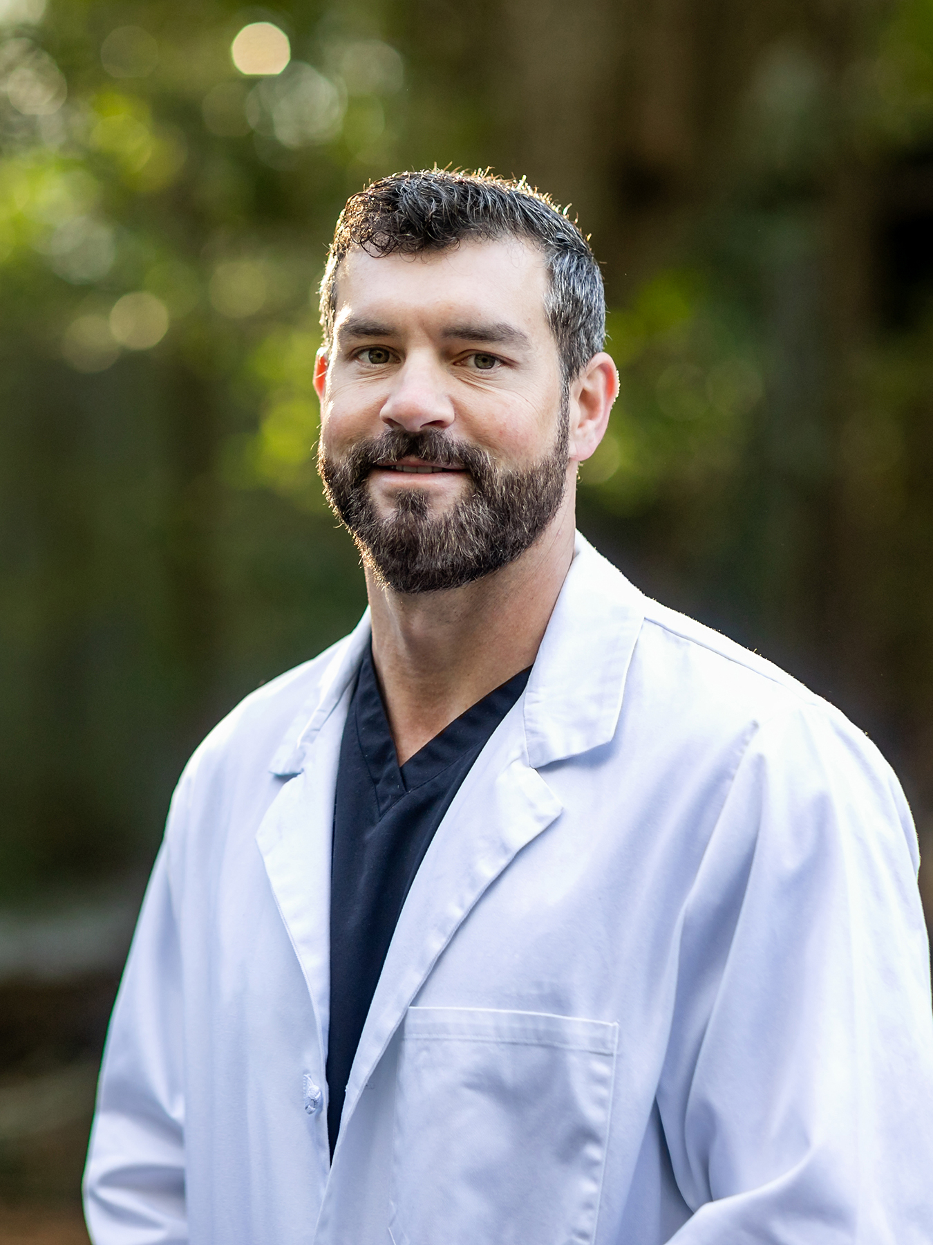 Image of a doctor with dark hair and a beard in a white lab coat standing outside in a natural green environment